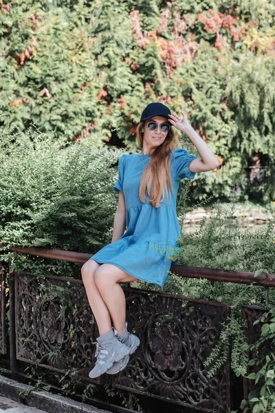 Улыбающаяся девушка в синем летнем платье сидит на металлических кованых перилах в летнем зеленом парке — стоковое фото