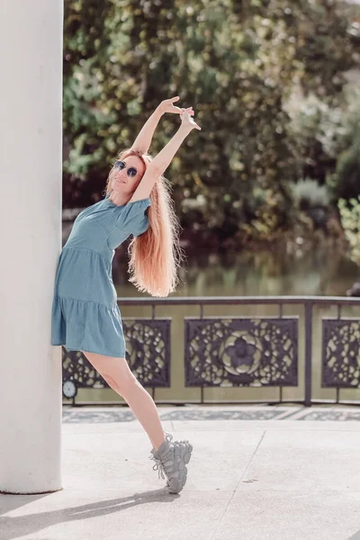 Una muchacha sonriente en un vestido corto está posando apoyada en una columna de pie sobre sus dedos de los pies y levantando sus manos contra el fondo de un estanque verde en el parque — Foto de Stock