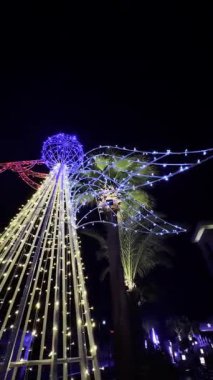 Aydınlatılmış melek çerçevesi, çelenkler ve palmiye ağacı ışığı gösterisiyle geceleyin.