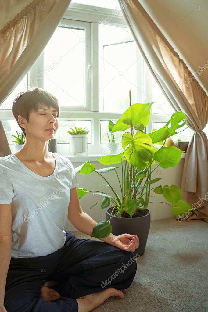 Woman practicing meditation sitting at cozy space with green plants near the window. Taking a break time for a minute, self-healing, and mindfulness. Mental health practice.