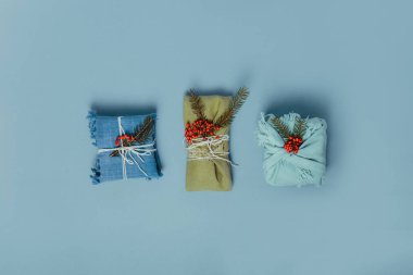 Sıfır noel ziyanı konsepti. Ladin dallı kumaşa sarılı üç hediye ve sessiz mavi arka planda böğürtlen. Flatlay. Minimalizm sürdürülebilir yaşam. Plastik yok. Boşluğu kopyala.