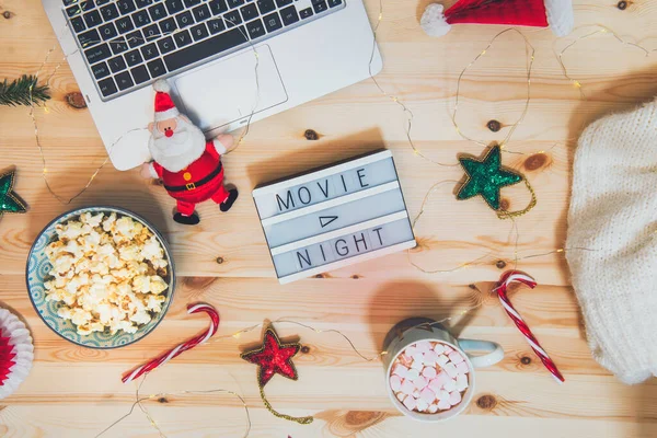Top view Christmas Movie night concept. Flat lay composition with Movie night message on the board, laptop, popcorn bowl, decor, a cup of cocoa with marshmallows and warm plaid on wooden background. — Stock Photo, Image