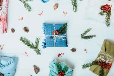 Sıfır noel ziyanı konsepti. Ladin dallı kumaşa sarılı hediye kutuları ve doğal malzemelerle beyaz ahşap arka planda kırmızı böğürtlenler. Flatlay. Sürdürülebilir yaşam. Plastik yok.