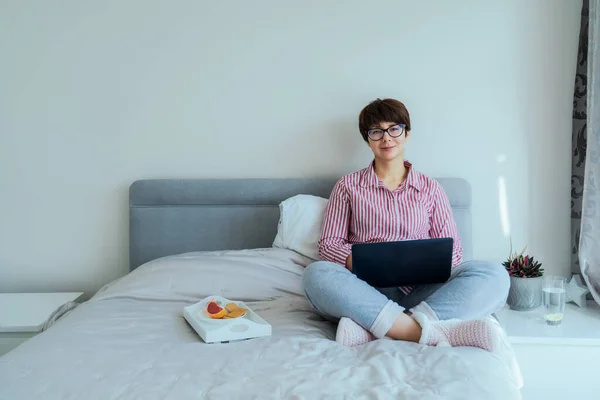 Mujer caucásica en gafas, camisa y pantalones de pijama usando portátil sentado en su cama y desayunando. Aspecto apropiado para el trabajo remoto desde el hogar durante el bloqueo. Enfoque selectivo, espacio de copia. — Foto de Stock