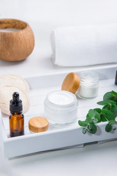 Herbal cosmetic creams in opened containers, fresh eucalyptus, loofah pads and cosmetic natural oil serum on a white wooden tray. Set for face self-care and DIY home eco-friendly hydration. Vertical. — Stock Photo, Image