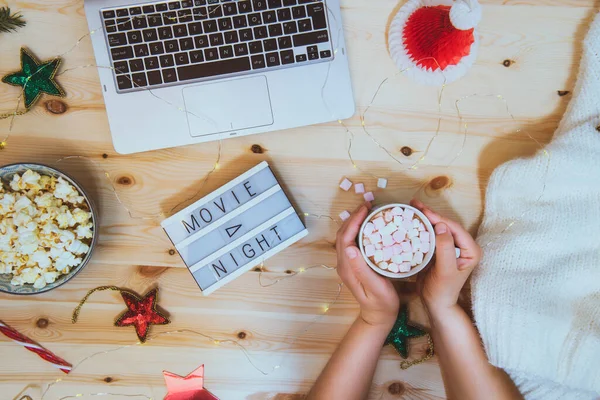 Top view woman holding cup of cococa with marshmellow and watching festive Christmas movie on laptop. Composition with Movie night message, popcorn bowl, decor, warm plaid on wooden background. — Stock Photo, Image