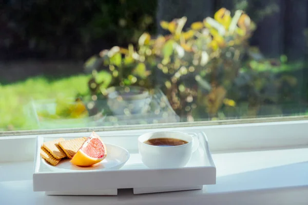 Tasse heißen Kaffee, Tee, Grapefruitscheibe und Kekse auf dem Tablett auf der Fensterbank, drinnen mit grünem, sonnigen Gartenhintergrund. Home Frühstück mit Aussicht. Selektiver Fokus, Kopierraum. — Stockfoto