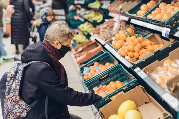 A young man with face covering mask selecting fruits in the store. Shopping during the pandemic. New normal lifestyle. Purchase food in grocery during coronavirus. Selective focus, copy space. — Stock Photo, Image