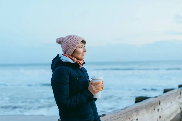 Smiling woman in a warm jacket and hat with reusable cup with hot drink standing on the pier and enjoying the winter sea view. Simple pleasures and local traveling, digital detox concepts. Copy space. — Stock Photo, Image