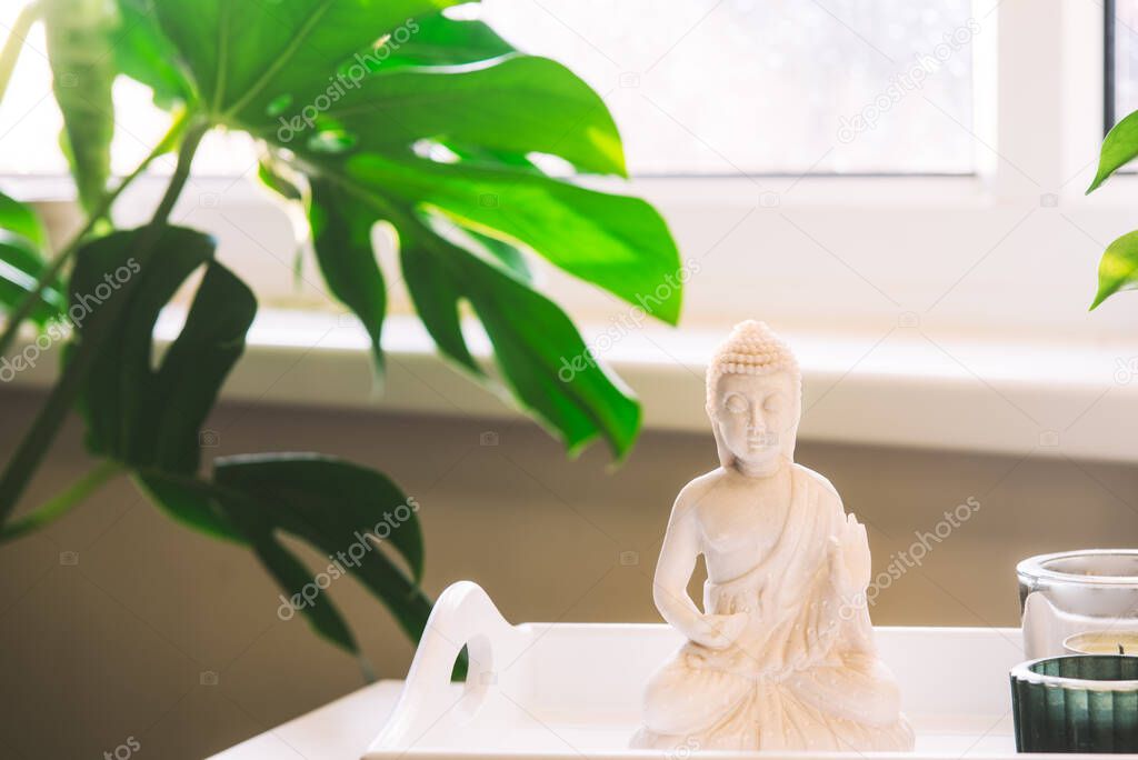 Decorative white Buddha statuette figure with candles standing on the tray near the window with a green monstera plant on the background. Meditation, relax ritual details. Selective focus copy space.