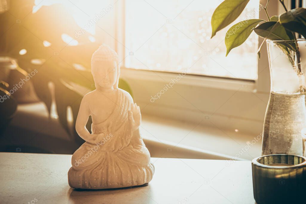 Decorative white Buddha statuette figure with candles standing on the tray near the window with a green monstera plant and bright sun light on the background. Meditation, relax ritual. Selective focus