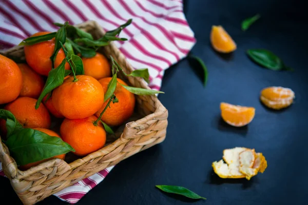 Färska mogna mandariner, klementin, mandarin med gröna blad i halmskålen på en randig servett och mörkblå sten bakgrund. Lokal citrusfruktskörd. Selektiv inriktning. — Stockfoto