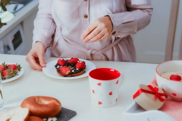 Frau verziert süßen Bagel mit Marmelade mit Erdbeeren und herzförmigen Bonbons. Vorbereitung des Überraschungsfrühstücks für Liebhaber am Valentinstag. Einfache Festtagsideen. Kopierraum — Stockfoto