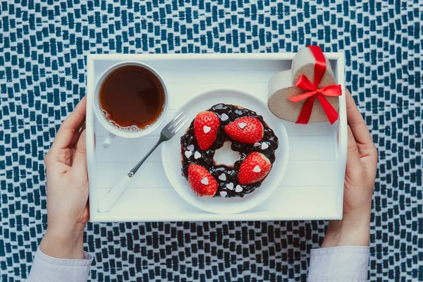 Frauenhände, die romantische Mahlzeiten für Liebhaber halten. Eine Tasse Tee, Bagel mit Marmelade mit Erdbeeren und herzförmigem Geschenkkarton auf dem weißen Holztablett. Einfache Überraschung zum Valentinstag — Stockfoto