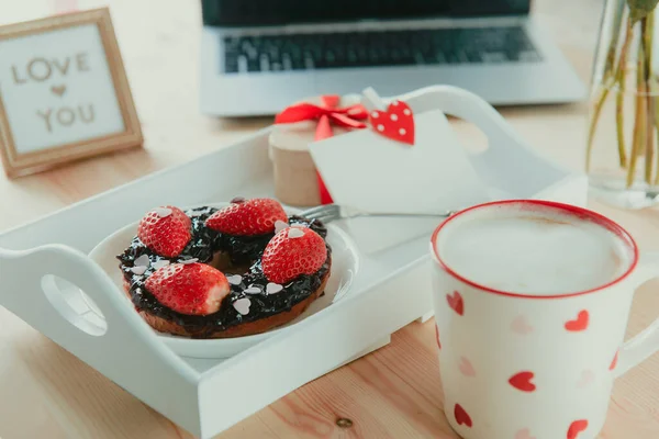 A tray with cup of latte coffee, bagel with strawberries and candy hearts, gift box and blank greeting card, Love you message on working place with laptop. Surprise for Valentine day. Selective focus. — Stock Photo, Image