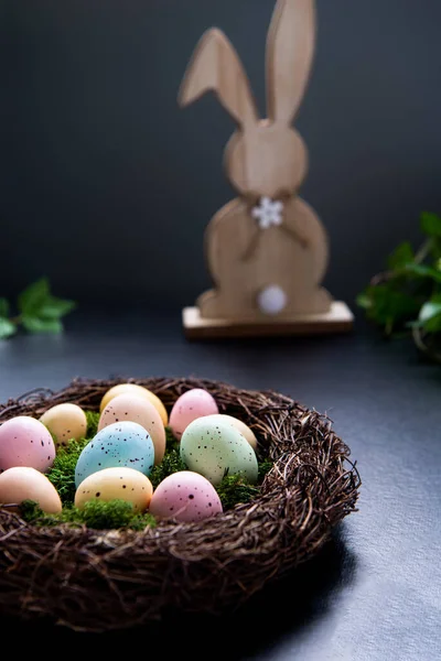 Ein Weidennest mit Moos und farbigen Ostereiern sowie einem hölzernen Hasen und einer grünen Pflanze auf dunklem Hintergrund. Frohe Osterfeiertage. Düstere Stimmung. Vertikale Karte. Selektiver Fokus. Kopierraum. — Stockfoto