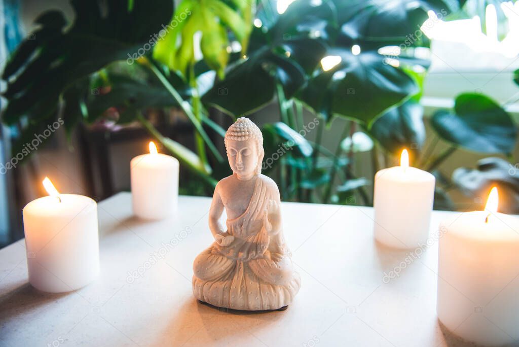Decorative white Buddha statuette with candles and green monstera plant on the background. Meditation and relaxation ritual. Exotic massage. Selective focus. Copy space.