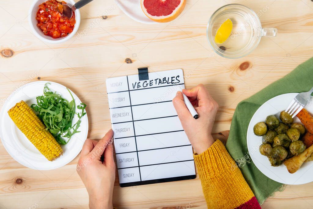 Diet planning and healthy meal. Top view Woman writing on board weekly vegetable menu. Ready meal portion of baked vegetables on the wooden table. New healthy lifestyle habits. Copy space.