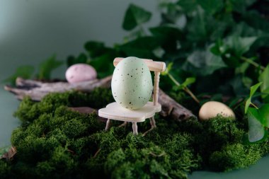 Paskalya yumurtasının yaratıcı bir kompozisyonu minyatür ahşap bir sandalyede sessiz koyu yeşil arka planda yosun ve ormanda duruyor. Peri masalı Paskalyası. Seçici odaklanma. Boşluğu kopyala.