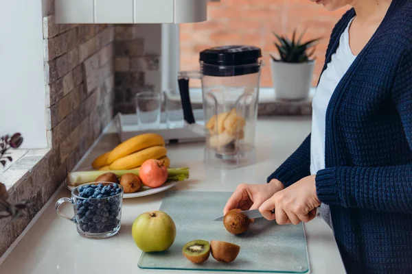 Vue de côté womans mains coupant kiwi frais pour préparer une boisson de désintoxication, smoothie aux fruits frais dans la cuisine à la maison. Une alimentation saine, végétarienne, végétalienne. Mise au point sélective, espace de copie. — Photo