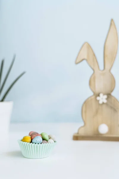 Pastel renkli paskalya şekeri yumurtaları bir kağıt kasede, ahşap tavşan ve açık beyaz ve mavi arka planda yeşil saksı bitkisi. Yumuşak seçici odaklanma. Paskalya minimalizmin kutlu olsun. Boşluğu kopyala. — Stok fotoğraf