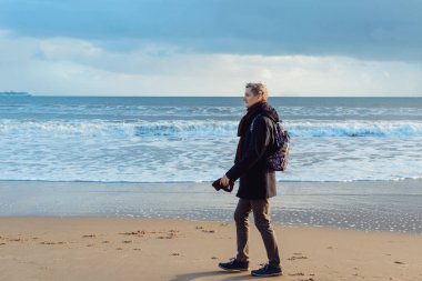 Sırt çantalı, kalın elbiseli genç bir adam kışın deniz kenarında yürüyor ve anın tadını çıkarıyor. Sahilde yürürken rahatla. Basit zevkler. Sonbaharda seyahat et, kışın. Seçici odaklanma. Boşluğu kopyala