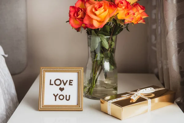 Caixa de presente dourada com fita, um buquê de flores de rosas em vaso e uma moldura com amor mensagem na mesa de cabeceira contra uma parede cinza. Dia dos namorados, aniversário, surpresa romântica do dia das mulheres. — Fotografia de Stock