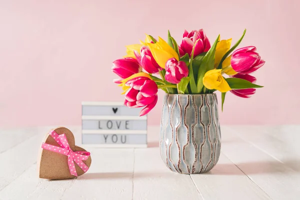 Recycled Heart shape gift box, spring bouquet of pink and yellow tulip flowers in a vase and Lightbox with love you message on the white wooden table and pink background. Greeting card mockup.