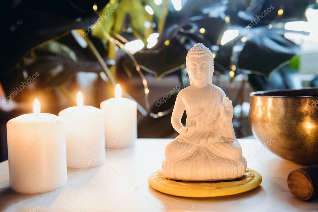Decorative white Buddha statuette , burning candles and singing bowl with green monstera plant on the background. Meditation and relaxation ritual. Exotic massage. Wide banner. Selective focus.