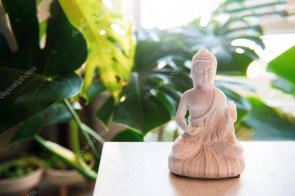 Decorative white Buddha statuette with candles and green monstera plant on the background. Meditation and relaxation ritual. Exotic massage. Selective focus. Copy space.