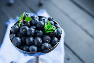 Mavi peçetede siyah ahşap arka planda nane yapraklarıyla süslenmiş antika metal kasede üst manzara yaban mersini. Karanlık ruh hali fotoğrafı. Dikey kart. Seçici odak, boşluğu kopyala