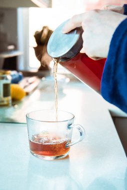 Evdeki bornozlu adam mutfak masasındaki fincana cam çaydanlıktan sıcak çay dolduruyor. Sabahları çay demleme süreci. Bir fincan taze demlenmiş siyah çay, ılık yumuşak ışık. Seçici odak