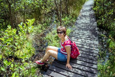Sırt çantalı bir kadın tahta yolda oturuyor ve yaz ormanında yürüyüş yaptıktan sonra dinleniyor. Tadını çıkarmak, doğayla bütünlük, dijital detoks, kişisel tatmin. İlkbaharda, yazda yerel seyahat.