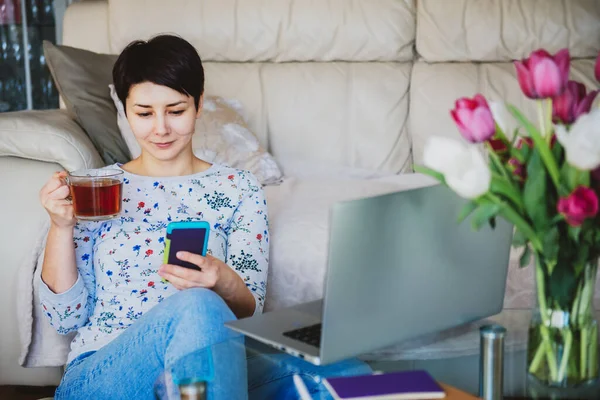 Kvinna rulla sociala nätverk på smartphone, chatta, tittar på video samtidigt koppla av och dricka te nära bekväm soffa hemma under pausen. Mysig modern arbetsplats med laptop. Kopiera utrymme. — Stockfoto
