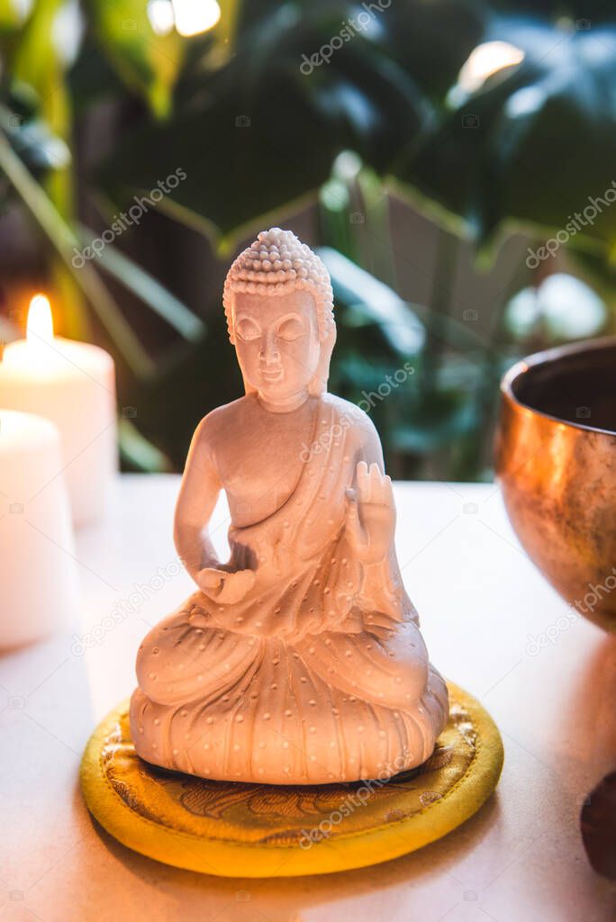 Decorative white Buddha statuette , burning candles and singing bowl with green monstera plant on the background. Meditation and relaxation ritual. Exotic massage. Vertical card. Selective focus