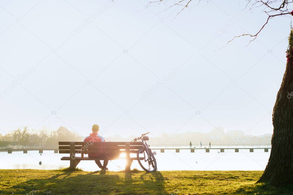 Back view man sitting on the bench at the bank of parks lake and enjoying the sunset. Enjoy nature, healthy break, digital detox. Selective focus, copy space.