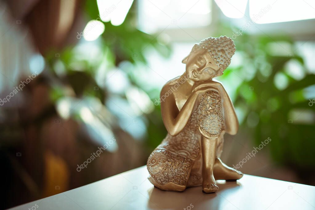 Decorative Golden bronze Buddha statuette with green monstera plant in room decor on the background. Meditation and relaxation ritual. Exotic massage. Selective focus. Copy space.