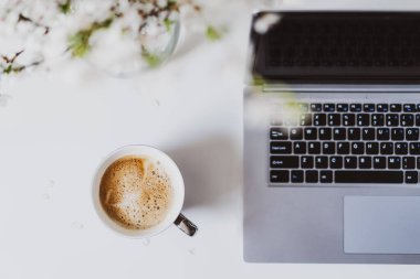 Top view workplace. White office table desk with a cup of fresh coffee, open laptop with keyboard and vase with blooming branches. Minimalist workspace. Freelance, online learning, home office.