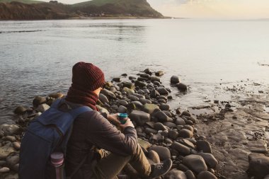 Taşın üzerinde oturan, sıcak çay ya da kahve içen sıcak giysili genç bir adam yeniden kullanılabilir termos bardaktan ve deniz manzarasından zevk alıyor. Sahilde yürürken rahatla. Sonbaharda, kışın kendi kendine seyahat eden yerel halk.
