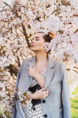 İlkbaharda, çiçek açan bahçede dikilen kapalı gözlü güzel romantik kadın. Gri montlu hassas genç manken. Dikey kart. Hassas bir ruh hali. Seçici odak, çekingen alan.