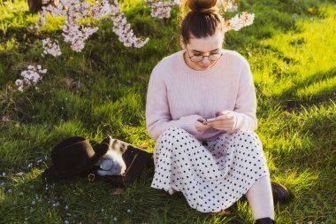 Beyaz tenli gözlüklü ve açık renk elbiseli genç bir kız parktaki çiçek açan ağacın yanındaki yeşil çimlerde oturuyor ve akıllı telefonunu kullanıyor. Kadın mesajlaşması, kayan sosyal ağlar, sohbet, video izleme.