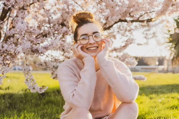 公園の花の木の近くの緑の芝生の上に座って眼鏡や薄いピンクの服で若い白人のブルネットのヒップスターの女の子の肖像画。Z世代の女の子は春の気分をお楽しみください。選択的焦点、コピースペース. — ストック写真