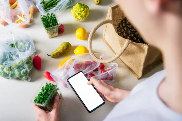 Online home food delivery of fresh vegetables and fruits. Young man holding phone and checking order list. Reusable bag with bio vegetables on white kitchen table. Local farmer healthy food.