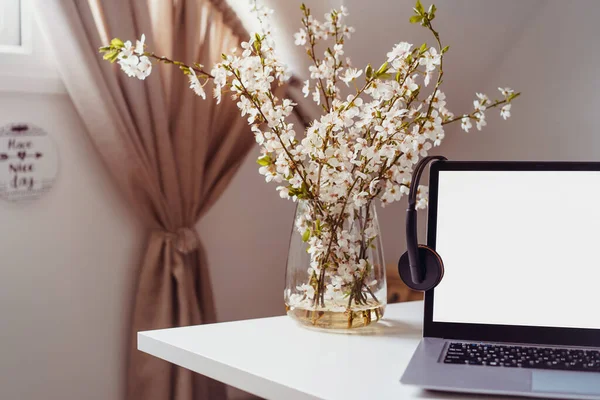 Um local de trabalho em casa com laptop de tela branca para maquetes, fones de ouvido e ramos floridos em um vaso na mesa com fundo interior do quarto. Freelance, trabalhando em casa, aprendizagem on-line, home office. — Fotografia de Stock