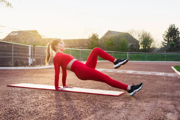 Mladá fitness žena v červeném sportovním oblečení dělá reverzní prkno koleno zastrčit cvičení na podložce. Cvičení venku na stadionu při západu slunce. Aktivní životní styl, sport a zdravotní péče. Kopírovat prostor — Stock fotografie