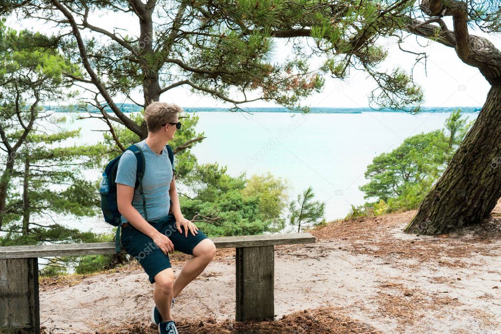 Man with backpack sitting on the bench and enjoying sea view during hiking walk in summer forest. Brownsea Island, England. Local Traveling and relax in nature. Digital detox Selective focus.