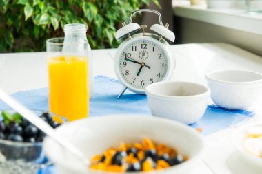 Kahvaltı zamanı. Sağlıklı yiyeceklerle birlikte çalar saat. Mısır gevreği, fındık ve böğürtlen, süt, mavi peçetede taze meyve suyu ve beyaz ahşap masa. Gündüz diyet planlaması ve sağlıklı yemek.