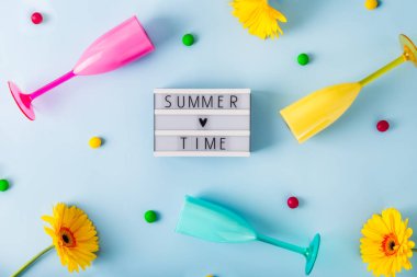 En üstteki düz ışıklı kutuda yazlık yazlık, parlak plastik kokteyl bardakları, çok renkli şeker damlaları ve açık mavi arkaplanda sarı gerbera çiçekleri. Yaz partisi havası.