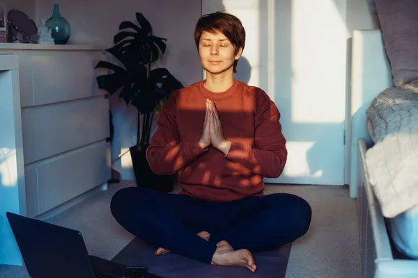 Uma mulher sentada em um tapete enquanto faz ioga posição Namaste no quarto. Relaxamento prática de ioga on-line em casa. Estilo de vida saudável. Treino online em casa. Treinamento online de fitness. Foco seletivo suave — Fotografia de Stock