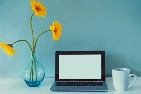 O local de trabalho moderno mesa com laptop mockup branco tela vazia, xícara de café, e flores de gerbera amarelo fresco no vaso no fundo azul. Espaço de trabalho no escritório em casa. Cor em design de interiores. — Fotografia de Stock